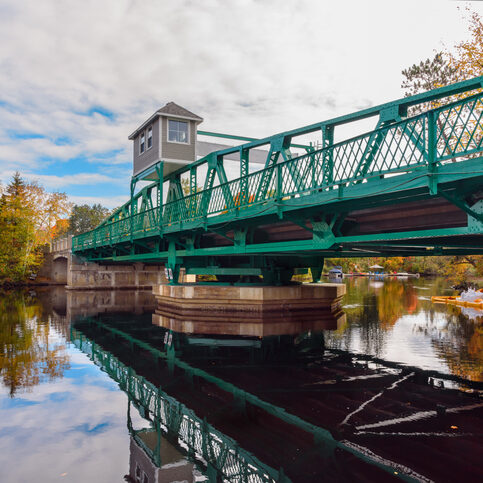 Road bridge over a river with wooded banks in autumn Swing bridge across a river on a partly cloudy autumn day. Reflection in water. Huntsville, ON, Canada.